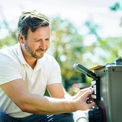 Man in a white polo shirt inspecting or repairing pool equipment outdoors.
