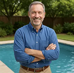 Smiling man in a blue shirt standing with arms crossed beside a backyard swimming pool.
