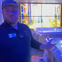 Man wearing a cap and black shirt presenting hot tubs in a showroom with bright lighting and scenic wall display.