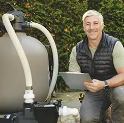 Smiling pool technician holding a clipboard beside pool filtration equipment outdoors.
