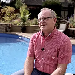 Smiling man wearing a red checkered shirt sitting beside a backyard swimming pool.