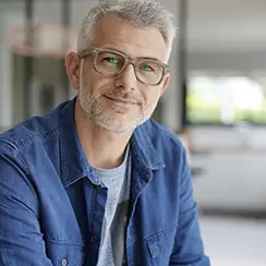 Smiling middle-aged man with gray hair and glasses wearing a denim shirt, sitting indoors.