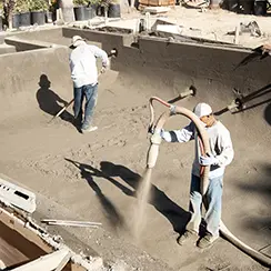 Construction workers applying concrete to form a swimming pool structure at a job site.